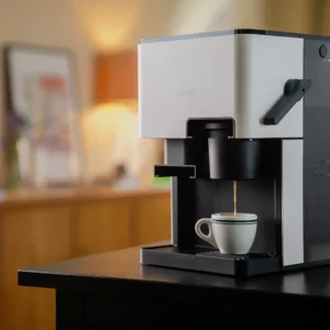 Ein NIVONA CUBE 4102 Kaffeeautomat in einem heimischen Ambiente brüht frischen Kaffee in eine weiße Espressotasse auf einem dunklen Holztisch.