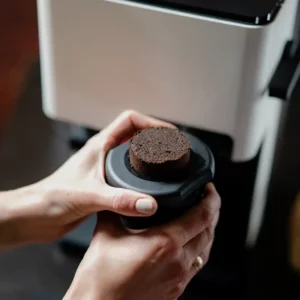 Hände, die einen kompakten Kaffeekuchen aus dem Click Cup des NIVONA CUBE 4102 Kaffeeautomaten entfernen, der eleganten Komfort und Effizienz veranschaulicht.