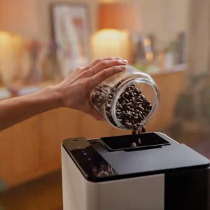 Eine Person füllt Kaffeebohnen in den Bohnenbehälter des NIVONA CUBE 4102 Kaffeeautomaten, der auf einer Küchenarbeitsplatte steht.
